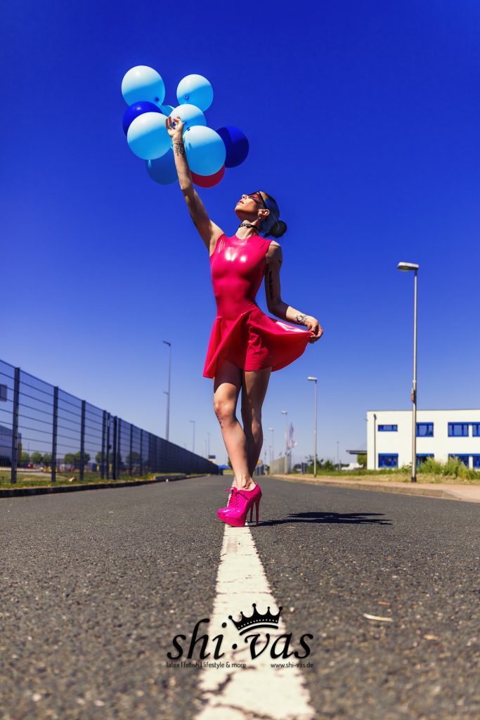 Coco im pinken Latexkleid blickt zu Luftballons nach oben auf einer Asphaltstraße