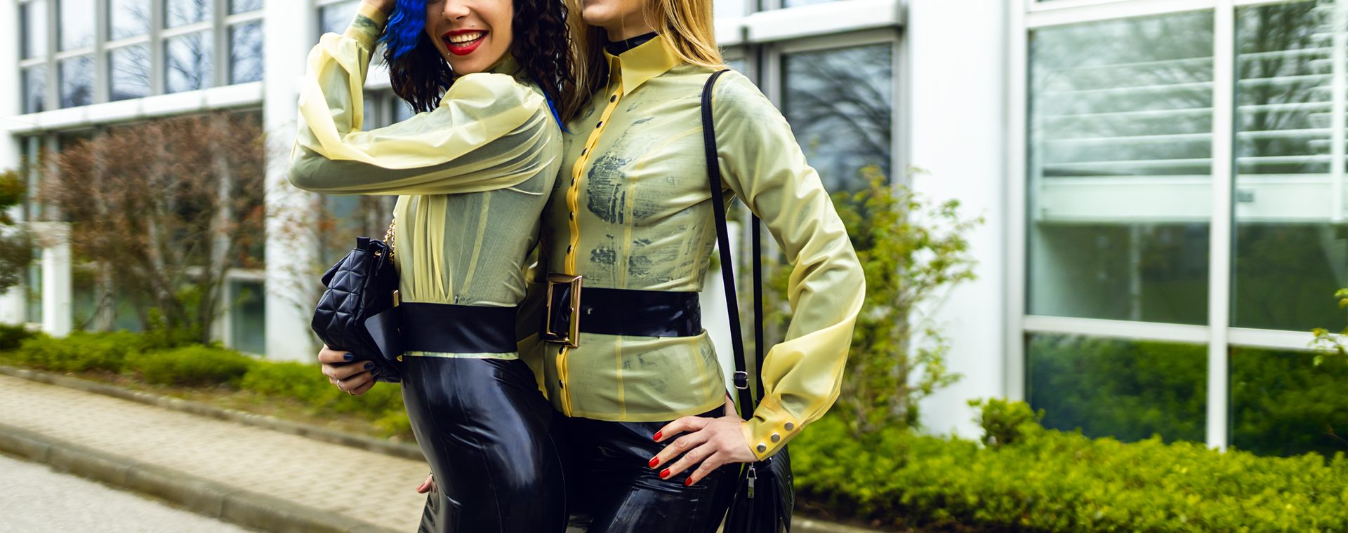 Zwei Frauen in identischen, hautengen Latex-Outfits (natur-transparente Blusen, schwarze Röcke, breite Gürtel) stehen eng aneinandergeschmiegt und lachen. Coco hat dunkles Haar mit blauen Strähnen, Lilly ist blond.