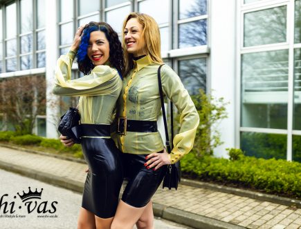 Zwei Frauen in identischen, hautengen Latex-Outfits (natur-transparente Blusen, schwarze Röcke, breite Gürtel) stehen eng aneinandergeschmiegt und lachen. Coco hat dunkles Haar mit blauen Strähnen, Lilly ist blond.