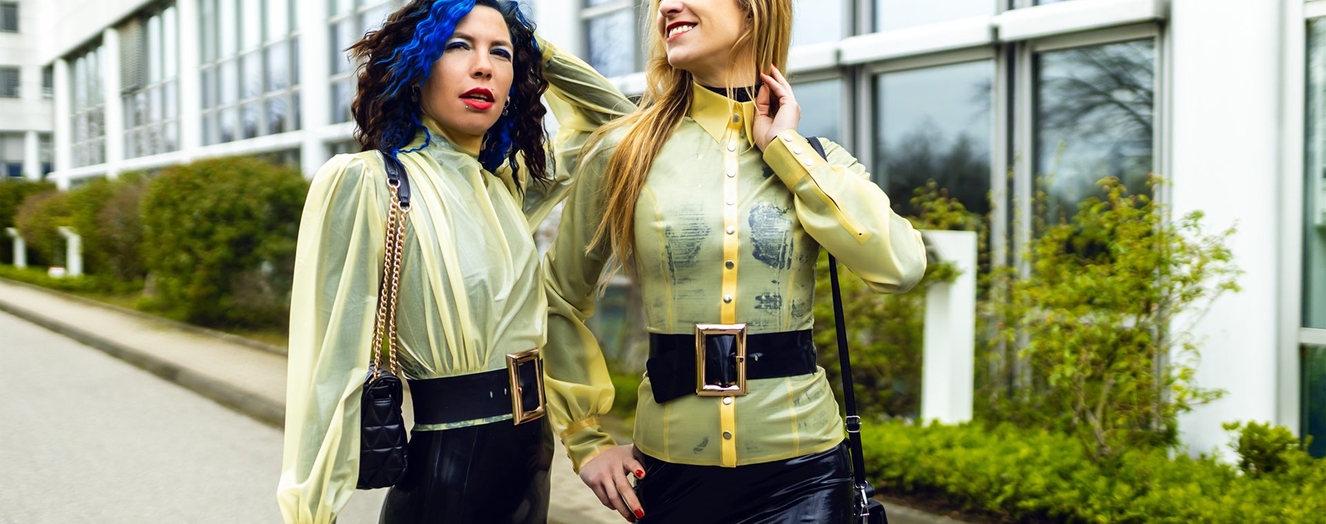 Coco und Lilly im identischen Latex-Look lächeln sich im Freien an.