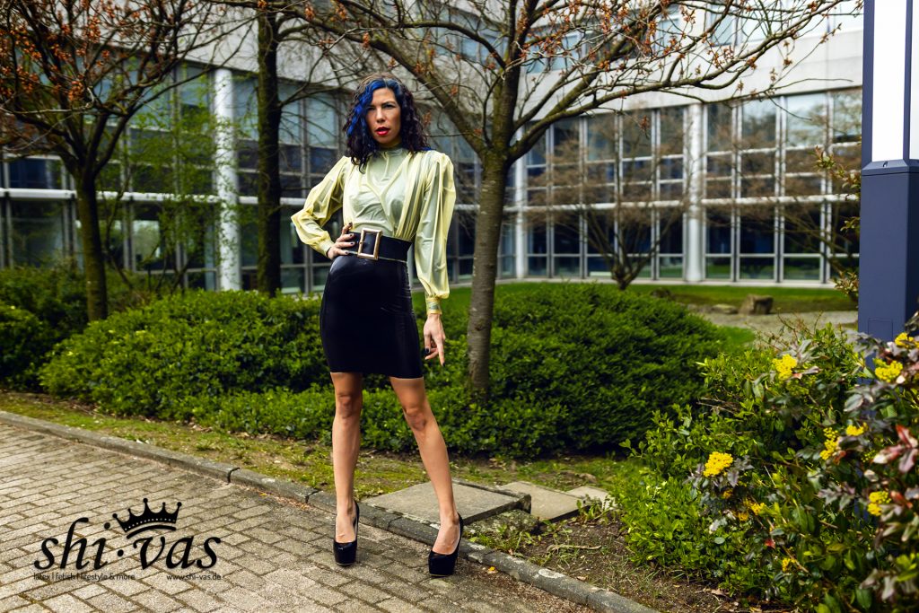 Coco blickt direkt in die Kamera, gekleidet in Latex-Bluse und Rock vor Sträuchern.