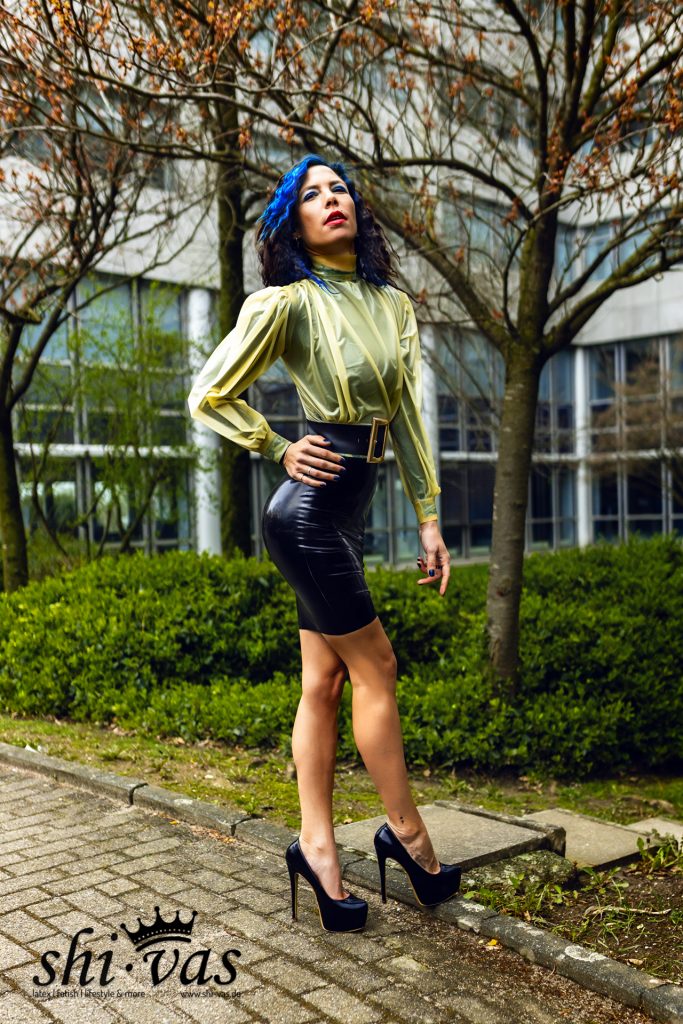 Coco mit stolzer Kopfhaltung, gelber Latexbluse und schwarzem Lackrock draußen.