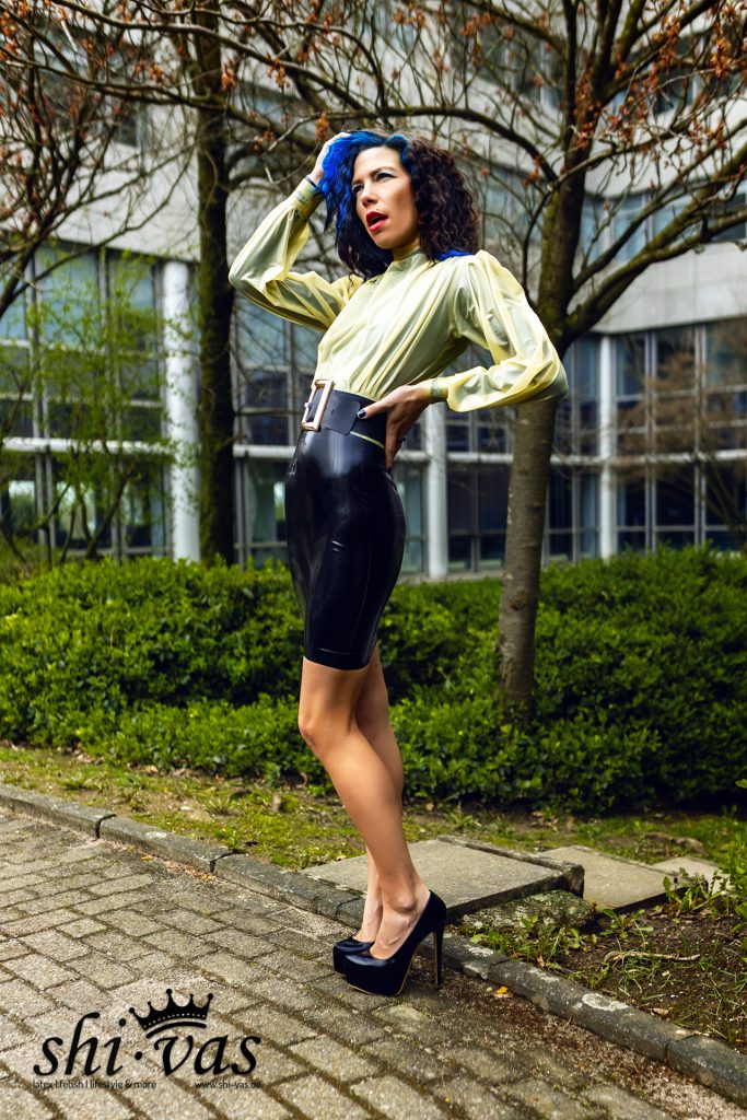 Coco posiert im eng anliegenden Latex-Outfit mit Hand am Kopf im Außenbereich.