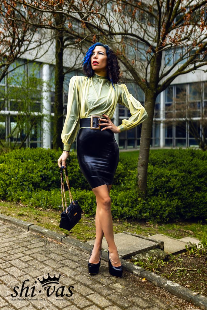 Coco mit schwarzer Handtasche, gelber Latexbluse und Lackrock in selbstbewusster Pose.