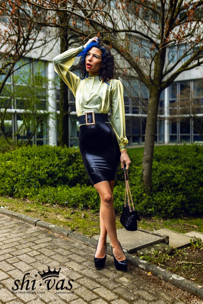Coco greift sich ins blaue Haar, trägt gelbes Latex-Oberteil und schwarzen Latexrock vor Gebäudekulisse.