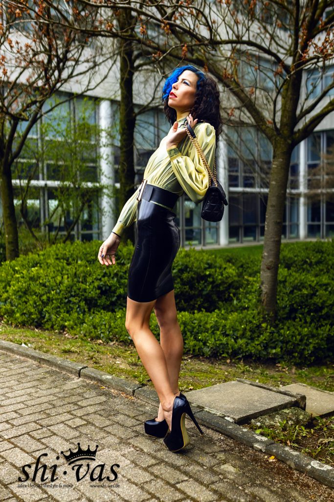 Coco im Profil mit gelber Latexbluse, schwarzem Latexrock und High-Heels im Freien.