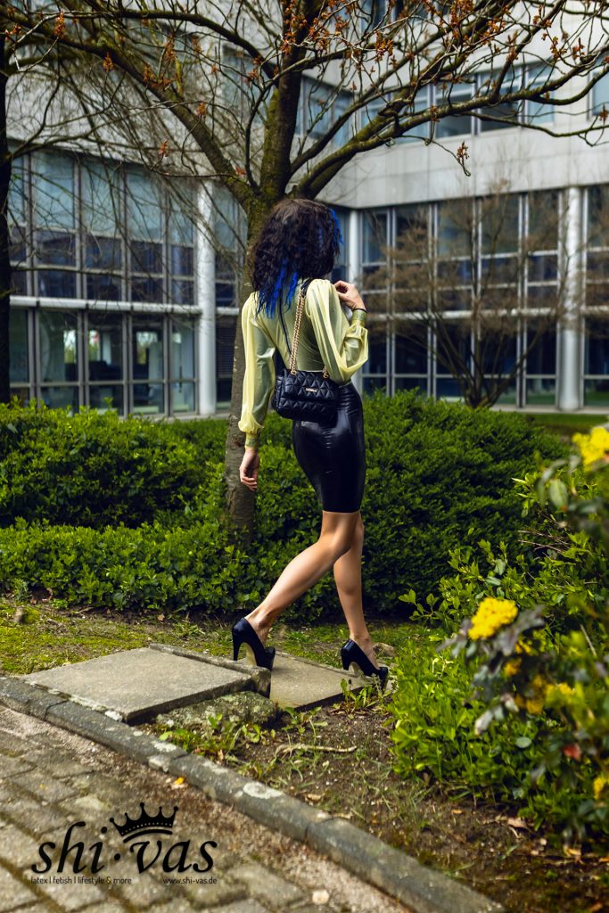Coco geht von der Kamera weg und blickt über die Schulter, getragenes Latex-Outfit vor Grünanlagen.
